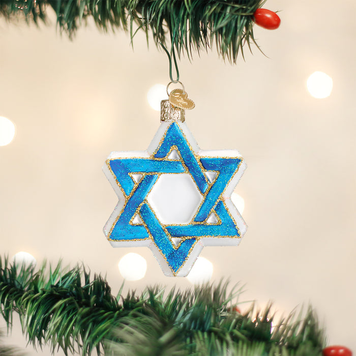 A blue and white Star of David ornament with gold accents hangs from a green Christmas tree branch, with blurred lights in the background.