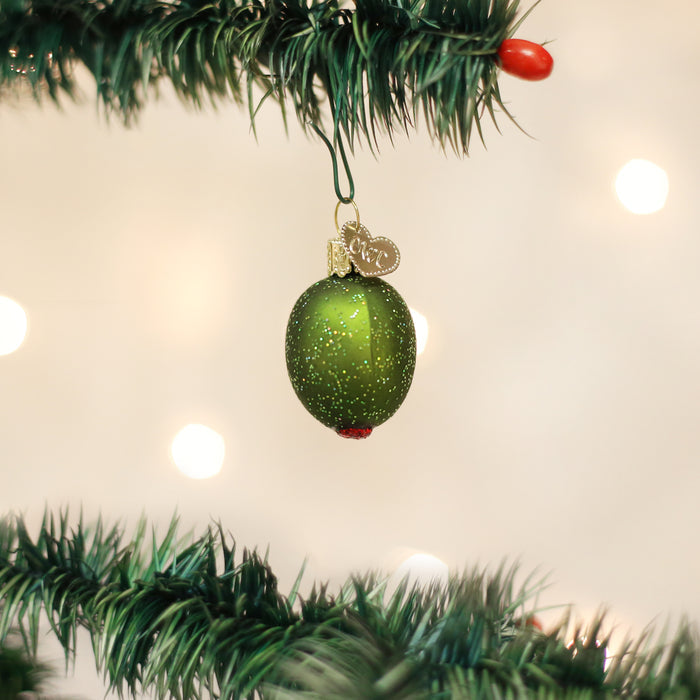 A green olive-shaped Christmas ornament with gold glitter and a red tip hangs from a Christmas tree branch; the background is softly lit with white bokeh lights.