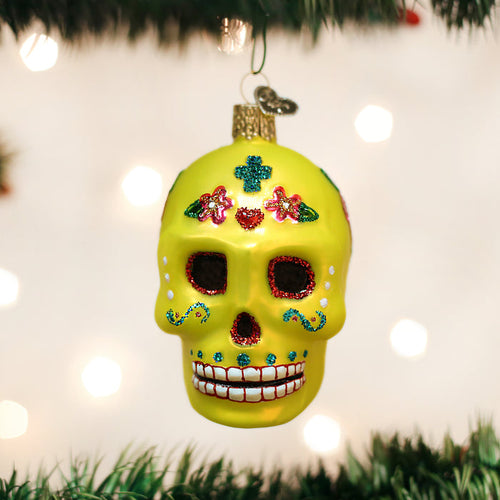 A bright yellow sugar skull ornament decorated with colorful glitter patterns hangs on a Christmas tree, with blurred lights glowing in the background.