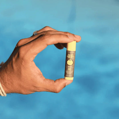 Hand holding Sun Bum Banana SPF 30 sunscreen lip balm against a blue background