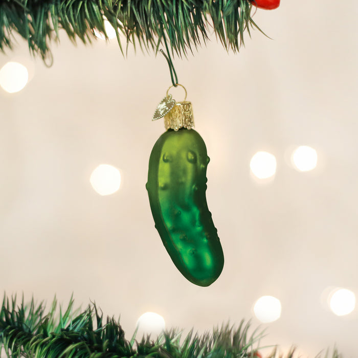 A green glass pickle ornament hangs from a Christmas tree branch with blurred white lights in the background.