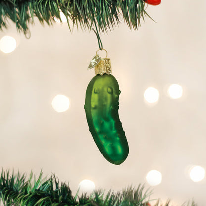 A green glass pickle ornament hangs from a Christmas tree branch with blurred white lights in the background.