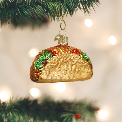 A Christmas tree ornament shaped like a taco hangs from a branch, surrounded by green pine needles and soft, blurred holiday lights in the background.