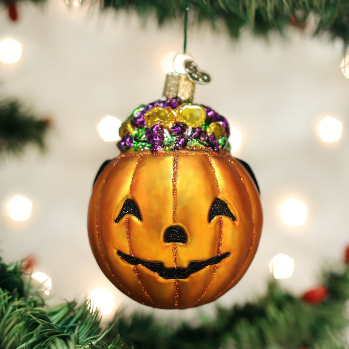 A shiny orange pumpkin ornament with a smiling face, decorated with colorful beads on top, hangs from a Christmas tree branch with blurred lights in the background.