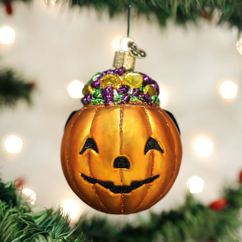 A shiny orange pumpkin ornament with a smiling face, decorated with colorful beads on top, hangs from a Christmas tree branch with blurred lights in the background.