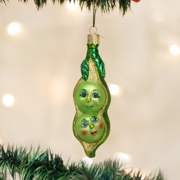 A Christmas tree ornament shaped like a pea pod with two smiling pea faces and glitter accents, hanging from a tree branch with blurred lights in the background.