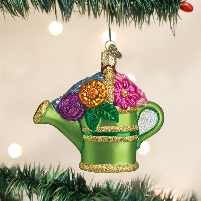 A colorful ornament shaped like a green watering can with gold glitter accents and vibrant flowers on top hangs from a Christmas tree branch, with white lights softly glowing in the background.
