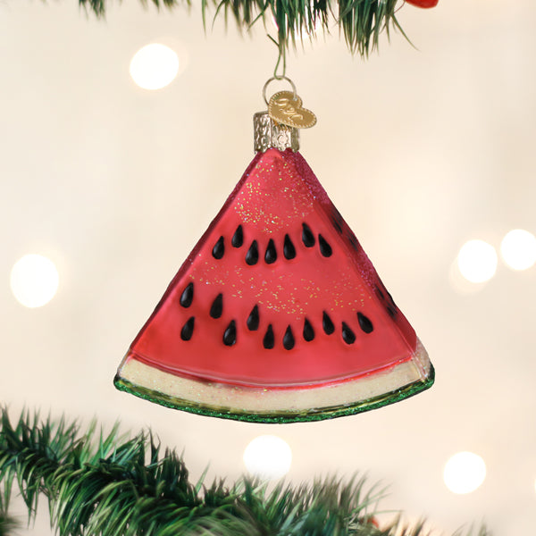 A Christmas tree ornament shaped like a slice of watermelon hangs from a branch, decorated with glitter and black seeds, with blurred lights in the background.