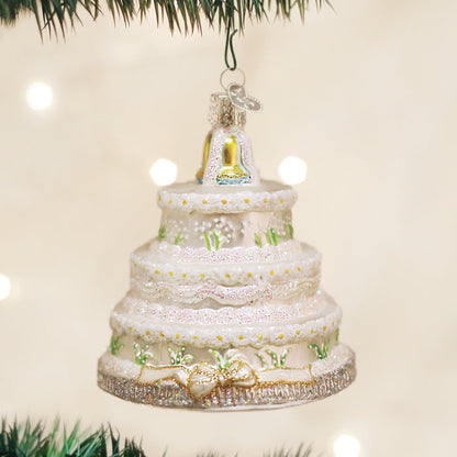A glass Christmas ornament shaped like a tiered white wedding cake with green accents, gold trim, and a gold bell topper, hanging from a tree branch with blurred lights in the background.