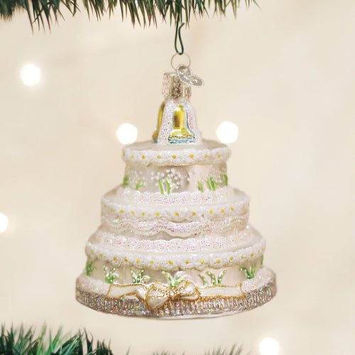 A glass Christmas ornament shaped like a tiered white wedding cake with green accents, gold trim, and a gold bell topper, hanging from a tree branch with blurred lights in the background.