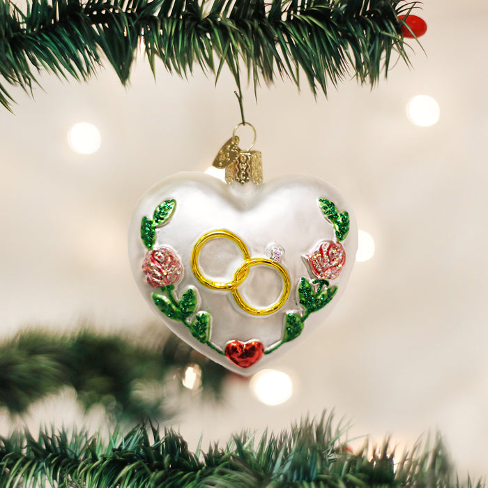 A heart-shaped Christmas ornament decorated with gold rings, a small gemstone, red roses, and green leaves hangs on a Christmas tree branch. White lights create a soft, festive background.