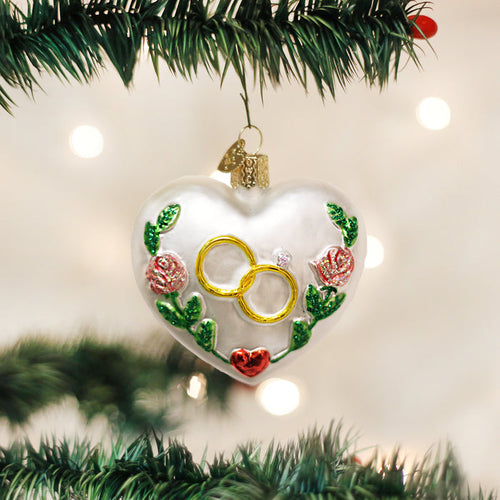 A heart-shaped Christmas ornament decorated with gold rings, a small gemstone, red roses, and green leaves hangs on a Christmas tree branch. White lights create a soft, festive background.
