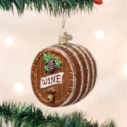 A Christmas tree ornament shaped like a wooden wine barrel with a grape cluster, a spigot, and the word WINE on the front, hanging on a green tree branch with soft lights in the background.